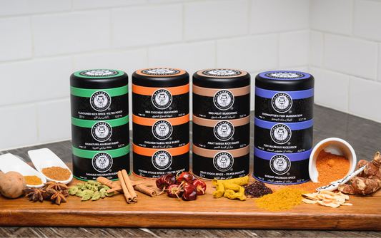 Four jars of Bahaurji spices on a wooden surface, each labeled with different types such as 'Rice Stack', 'Meat Stack', 'Chicken Stock', and 'Fish Stack', accompanied by an array of visible spices and seasonings.
